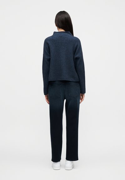 Στρατιωτικό πουλόβερ με ψηλό γιακά και φαρδιά εφαρμογή, σε συνδυασμό με σκούρο τζιν παντελόνι. Υφή υφάσματος με ευθεία γραμμή στα πόδια. Λευκά αθλητικά παπούτσια.