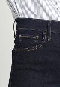 Calças de ganga de denim azul-escuro com cintura alta, apresentando costuras amarelas, um pequeno bolso dianteiro e fecho com botão.