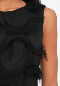 Primer plano de un vestido negro sin mangas con grandes flecos circulares texturizados en la parte superior del torso y el brazo de una persona.