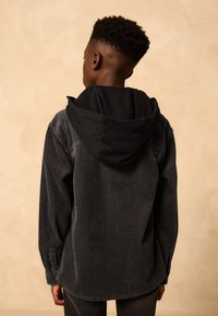 Black corduroy jacket with a hood, featuring ribbed texture, long sleeves, and button cuffs. Seen from the back against a neutral background.