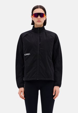 Frau trägt schwarze Jacke mit Reißverschluss und schwarze Leggings, mit kurzen dunklen Haaren und reflektierenden Sportsonnenbrillen, steht vor einem schlichten Hintergrund.