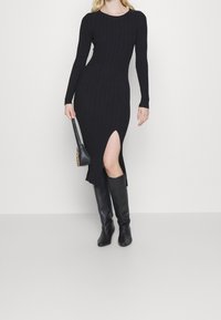 Vestito nero a costine con maniche lunghe e uno spacco laterale alto fino alla coscia. Abbinato a stivali neri al ginocchio e a una piccola borsa.