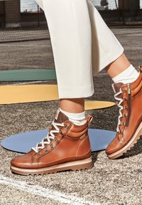 Brown leather ankle boots with white laces and a side zipper, featuring a textured upper, a chunky sole, and a padded collar.