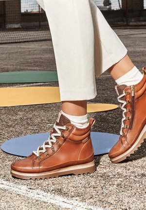 Bottines en cuir marron avec des lacets blancs et une fermeture éclair sur le côté, présentant un dessus texturé, une semelle épaisse et un col rembourré.