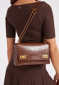 Donna che indossa un vestito marrone a coste, mentre porta una borsa a spalla Guess marrone con texture, dotata di catene dorate e dettagli in oro.