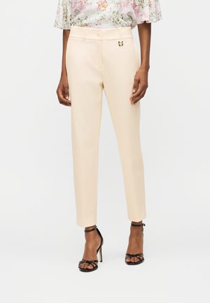 Femme portant un pantalon beige longueur cheville, un chemisier floral avec des manches à volants, et des sandales à talons hauts noires à brides autour de la cheville.