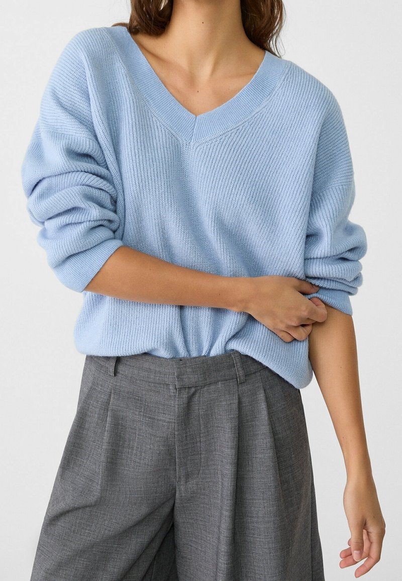 Donna che indossa un maglione azzurro chiaro con scollo a V e maniche arrotolate, infilato in pantaloni grigi a pieghe a vita alta, su uno sfondo neutro.