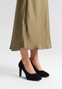 Czarne zamszowe szpilki z okrągłym noskiem i gładką teksturą, zestawione z zwiewną oliwkowozieloną satynową spódnicą.