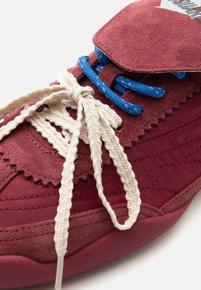 Gros plan sur une basket en daim bordeaux avec des œillets bleus et des lacets épais blancs noués, sur une section supérieure en tissu matelassé.