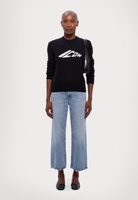 Zwarte gebreide trui met wit logo, gecombineerd met lichtblauwe cropped jeans en zwarte ballerina's. Eenvoudig ontwerp, rechte pasvorm, geribbelde manchetten.
