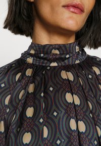 Monivärinen paita, joka on valmistettu sileästä kankaasta ja jossa on korkea kaulus, pleatattu muotoilu sekä geometrinen kuvio tummissa ja vaaleissa sävyissä.