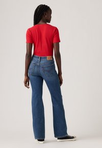 Red ribbed short-sleeve top paired with high-waisted flared blue denim jeans. Notable back pocket design with brand patch. Black sneakers.