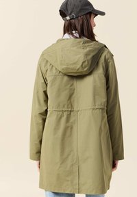 Olivgrön parkacoat med huva, med en åtdragen midja, mjuk textur och rak skärning, visad bakifrån mot en neutral bakgrund.