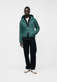 Άνδρας που φοράει πράσινο παλτό με κουκούλα, μαύρο πουκάμισο, μαύρο παντελόνι και λευκά αθλητικά παπούτσια, στέκεται με το ένα χέρι στην τσέπη του παλτού.