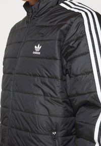 Zwarte donzen jas met horizontale doorgestikte banen, wit Adidas-logo op de borst en witte drie-streep accenten langs de mouwen. Voorzien van een ritssluiting op de zak.
