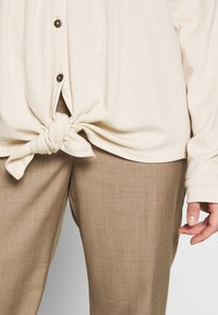 Beige, lätt stickad topp med knut framtill; har bruna knappar och långa ärmar. Matchad med strukturerade, ljusbruna byxor.