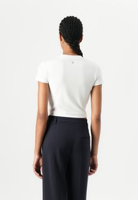 Hvit kortermet cropped skjorte, glatt stoff, rund hals, logo detaljer bak, kombinert med skreddersydde mørke bukser, vist bakfra.