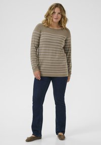 Femme debout portant un pull beige à rayures, un jean bleu foncé et des ballerines marron, devant un fond clair.
