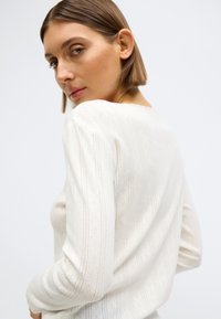 Πλεκτό τοπ σε κρεμ απόχρωση με περίπλοκο μοτίβο και κάθετες ρίγες. Ελαφρύ ύφασμα με υφή και μακριά μανίκια.