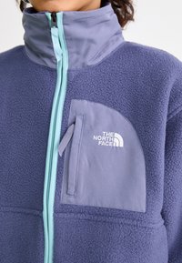 Fioletowa polarowa kurtka z wysokim kołnierzem, niebieskim zamkiem błyskawicznym i kieszenią na lewym piersi. Posiada białe logo "The North Face". Miękka w dotyku.