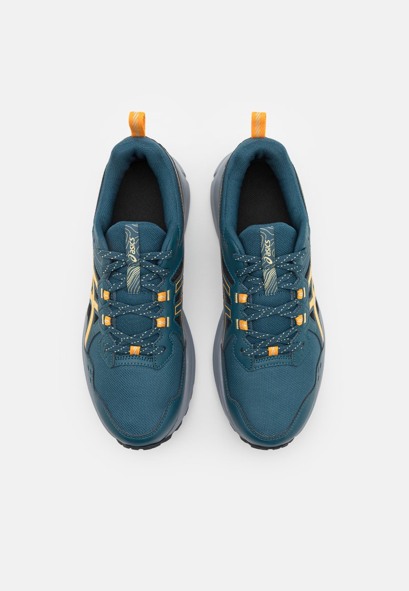 Baskets de sport teal avec des accents noirs et dorés, présentant une tige texturée, des lacets ronds et un onglet de talon orange. Semelle en caoutchouc.