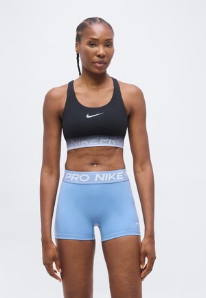 Femme debout portant un soutien-gorge de sport Nike noir et un short de sport Nike Pro bleu clair sur un fond blanc uni.