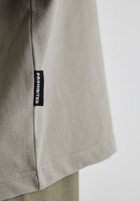 Tricou din bumbac gri, cu o margine dreaptă, dotat cu un eticheta neagră pe care este scris cu alb "PROHIBIT" pe cusătura laterală.