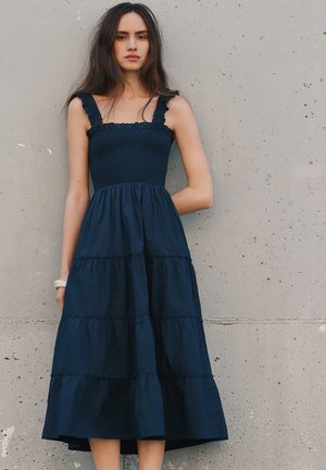 Jeune femme aux longs cheveux foncés portant une robe sans manches bleu foncé à volants, debout contre un mur en béton gris.