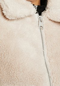 Luddig beige jacka med dragkedja framtill, med krage och silverfärgad dragkedja. Texturen verkar mjuk och plyschig.