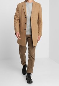 Pískový kabát s přiléhavým střihem, s třemi černými knoflíky, nošený přes světle šedý svetr s pleteným vzorem a hnědé kalhoty. Černé boty dotvářejí celý outfit.