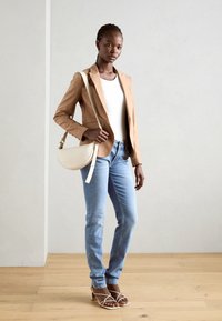 Světle hnědý blazer přes bílý top, doplněný světle modrými džíny. Béžová kabelka na rameno a bílé sandály na podpatku dotvářejí celý outfit.