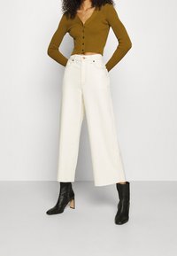 Avkortad olivgrön ribbad cardigan med knäppning, i kombination med högmidjade krämfärgade vida byxor. Svarta ankelskors med klack.