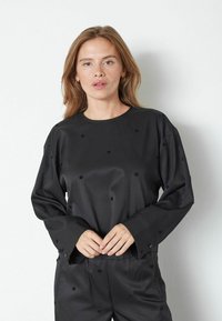 Top lungo in satin nero a maniche lunghe con scollo rotondo, caratterizzato da pois ricamati e una vestibilità rilassata. Dettagli con bottoni sulle maniche.