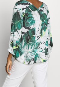 Lekka, zwiewna bluzka z tropikalnym wzorem liści w odcieniach zieleni, bieli i czerni. Posiada dekolt w kształcie litery V oraz elastyczne rękawy.