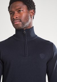 Granatowy sweter z dzianiny z prążkowanym, wysokim kołnierzem, zamkiem na ćwiartkę oraz małym logo na piersi. Gładka faktura, dopasowany krój.