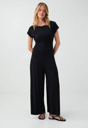 Femme aux longs cheveux blonds portant une combinaison noire à manches courtes et des sandales noires, debout devant un fond clair uni.