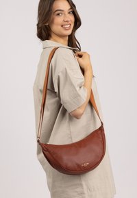 Braune Leder-Schultertasche mit runder Form, verstellbarem Tragegurt und einem dezenten Logo. Glatte Textur und minimalistisches Design.