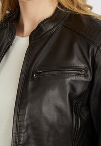 Giubbotto di pelle nera con colletto rotondo, dotato di tasca sul petto con zip e spalle imbottite. Texture liscia e design aderente.