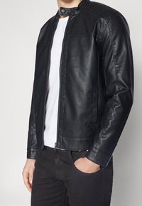 Veste en cuir noir avec un col montant et une fermeture éclair à l'avant. Elle présente une texture lisse et deux poches latérales. Portée par-dessus une chemise blanche.