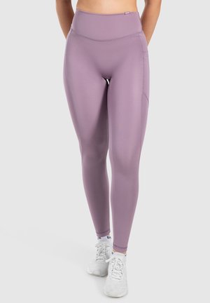 Leggings lavanda a vita alta realizzati in tessuto elastico, con una texture liscia e dettagli di tasche laterali, indossati con scarpe sportive bianche.