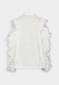 Blouse blanche avec broderie florale colorée, dotée de manches volantes et de bords festonnés. Texture légère et douce avec une coupe ample.