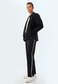 Sudadera negra con cremallera y bolsillos, combinada con pantalones negros que tienen rayas laterales blancas y zapatillas blancas. Tejido suave, corte relajado.