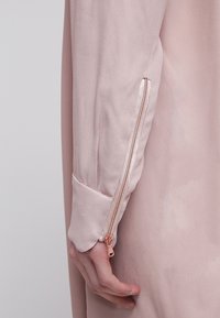 Vêtement à manches longues rose clair avec une fermeture éclair or rose au poignet, porté par une personne avec la main détendue le long du corps.