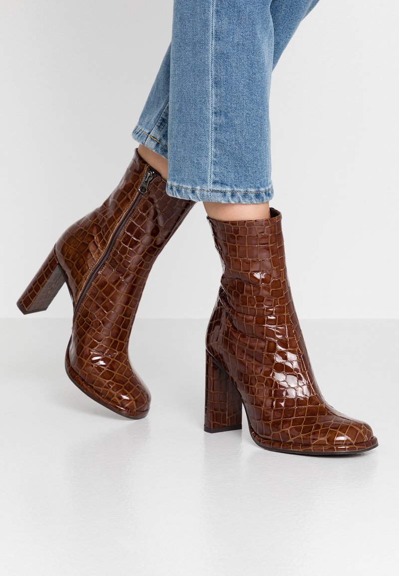 Bottines en cuir verni marron avec une texture crocodile, dotées d'un talon bloc haut et d'une fermeture éclair latérale, portées avec un jean bleu raccourci.