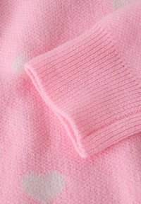 Roza pletena pulover podrobnost z mehkoba, rebrastimi manšeti in subtilnimi belimi srčnimi vzorci. Poudarek je na tkanini in oblikovalskih elementih.