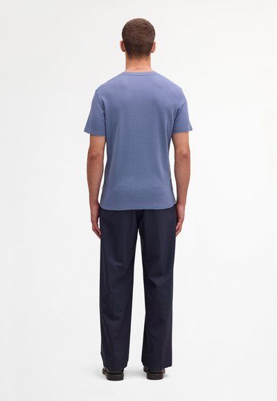 Uomo in piedi di spalle che indossa una camicia azzurro chiaro a maniche corte, pantaloni blu scuro e scarpe nere su uno sfondo bianco uniforme.