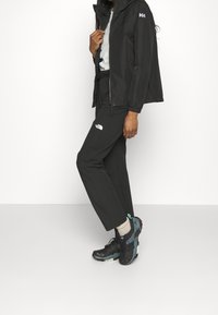 Svart vattentålig jacka med justerbar huva, kombinerad med svarta softshell-byxor med en logotyp och sportiga svarta och teal skor.