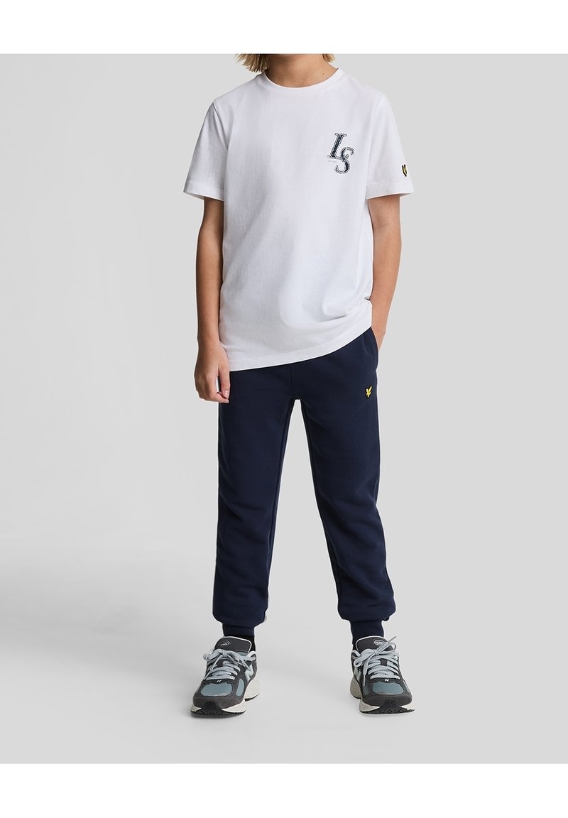 Witte katoenen T-shirt met "LS" logo op de borst, gecombineerd met marineblauwe fleece joggers. Grijze sportieve schoenen met blauwe accenten. Eenvoudig ontwerp.