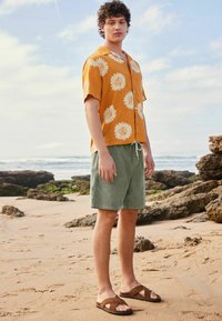 Korte mouwen, oranje shirt met zonnebloemprint, gecombineerd met groene shorts en bruine sandalen, tegen een strandachtergrond met rotsen.