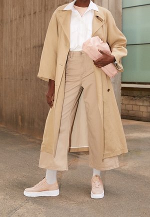 Beige trench coat, valkoinen paita, vaaleanbeige leveälahkeiset housut, vaaleanpunaiset tossut valkoisilla pohjilla, pitäen vaaleanpunaista clutch-laukkua. Sileä kangas, hyvin muotoiltu design.